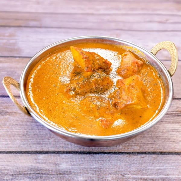 A metal karahi bowl filled with vibrant orange Indian curry and paneer, garnished with herbs.